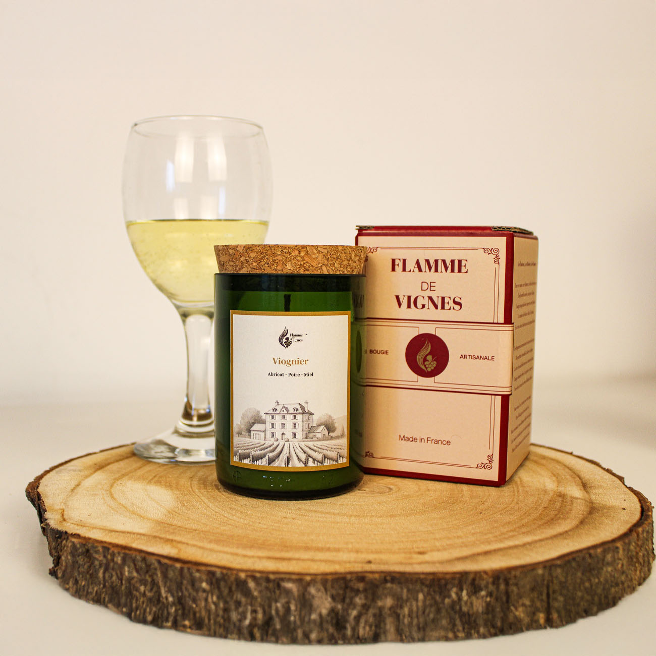 Bougie parfumée artisanale faite main Viognier, posée sur une tranche de bois avec sa boîte et un verre de vin blanc.