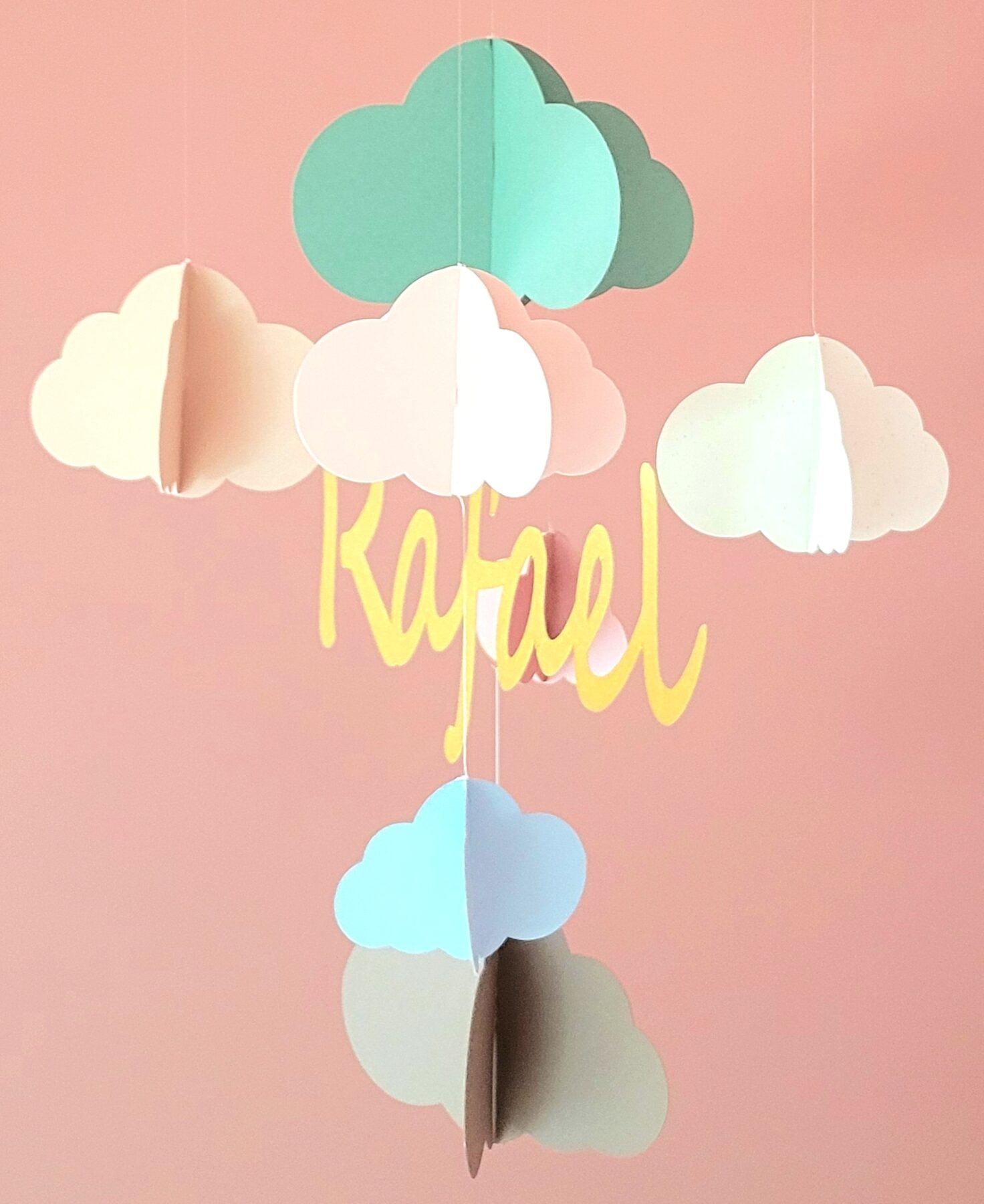 Personnalisable avec le prénom de votre choix, il est suspendu à un cercle en bois et agrémenté de nuages en 3D dans des tons pastel harmonieux.