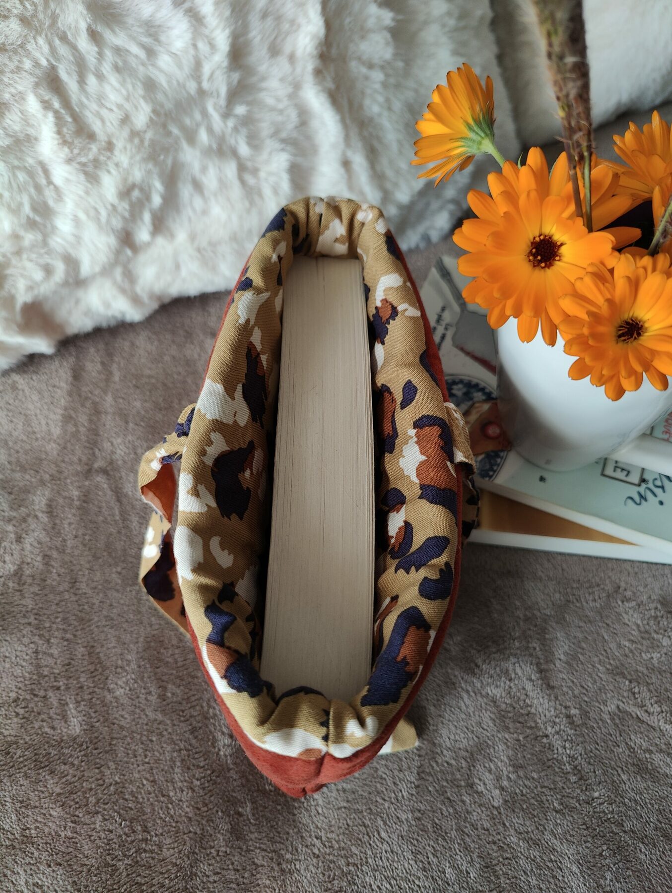 Intérieur tacheté d’une pochette en tissu couleur rouille contenant un livre, posée sur un lit avec oreillers.