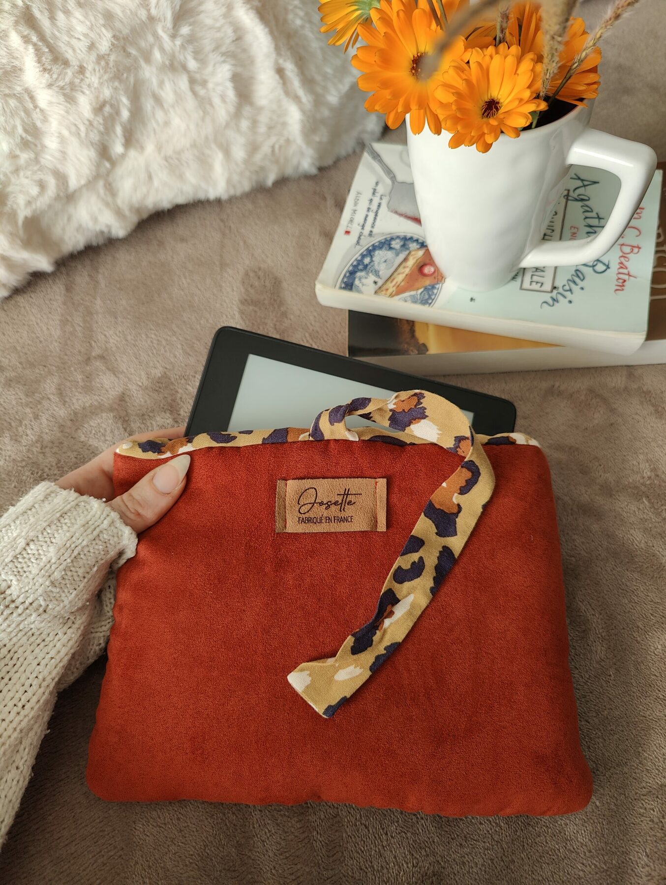 Pochette couleur rouille avec intérieur tacheté contenant une liseuse, posée sur un lit avec oreillers.