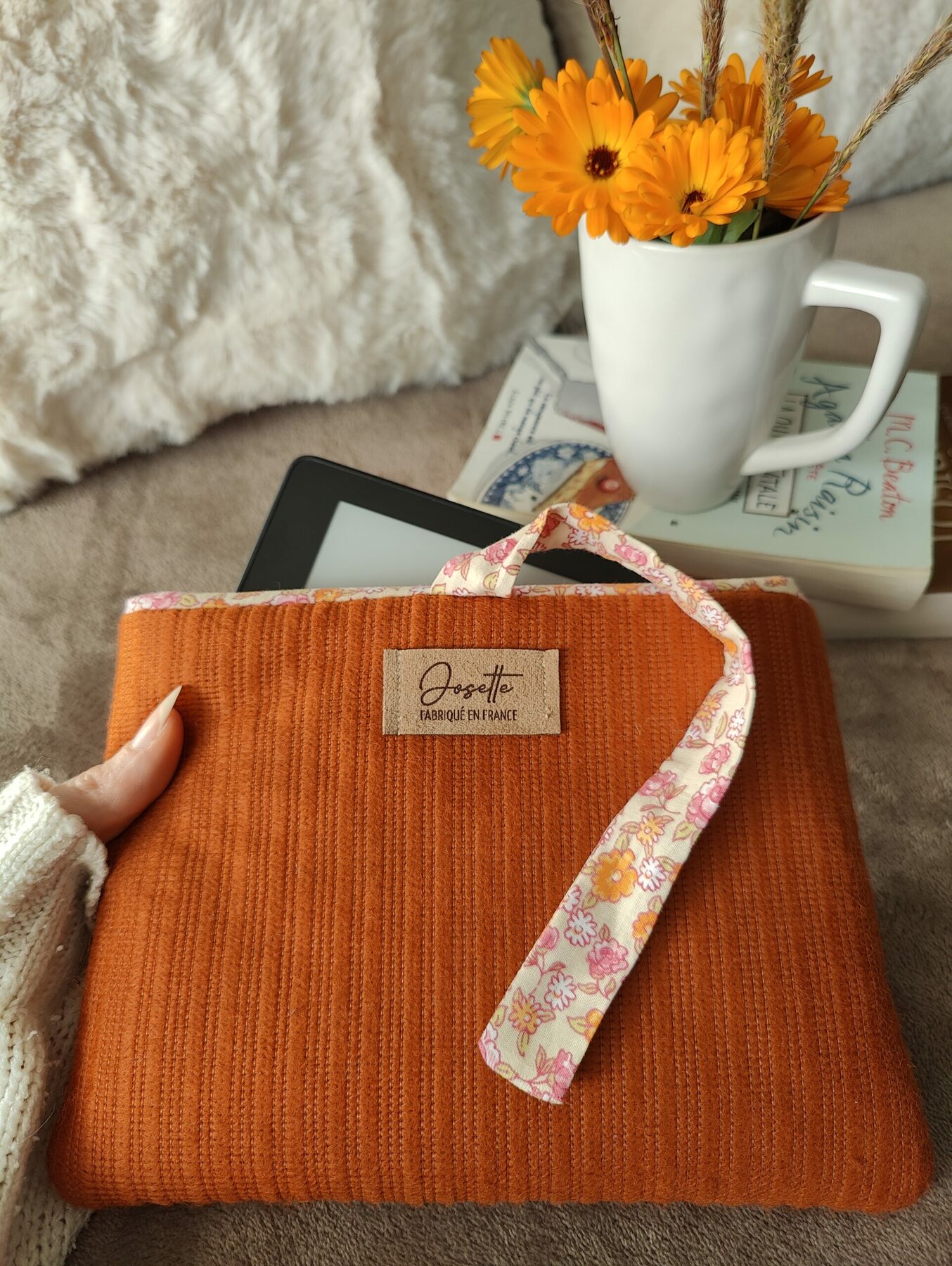 Pochette en tissu orangé avec intérieur fleuri contenant une liseuse, posée sur un lit avec deux oreillers.