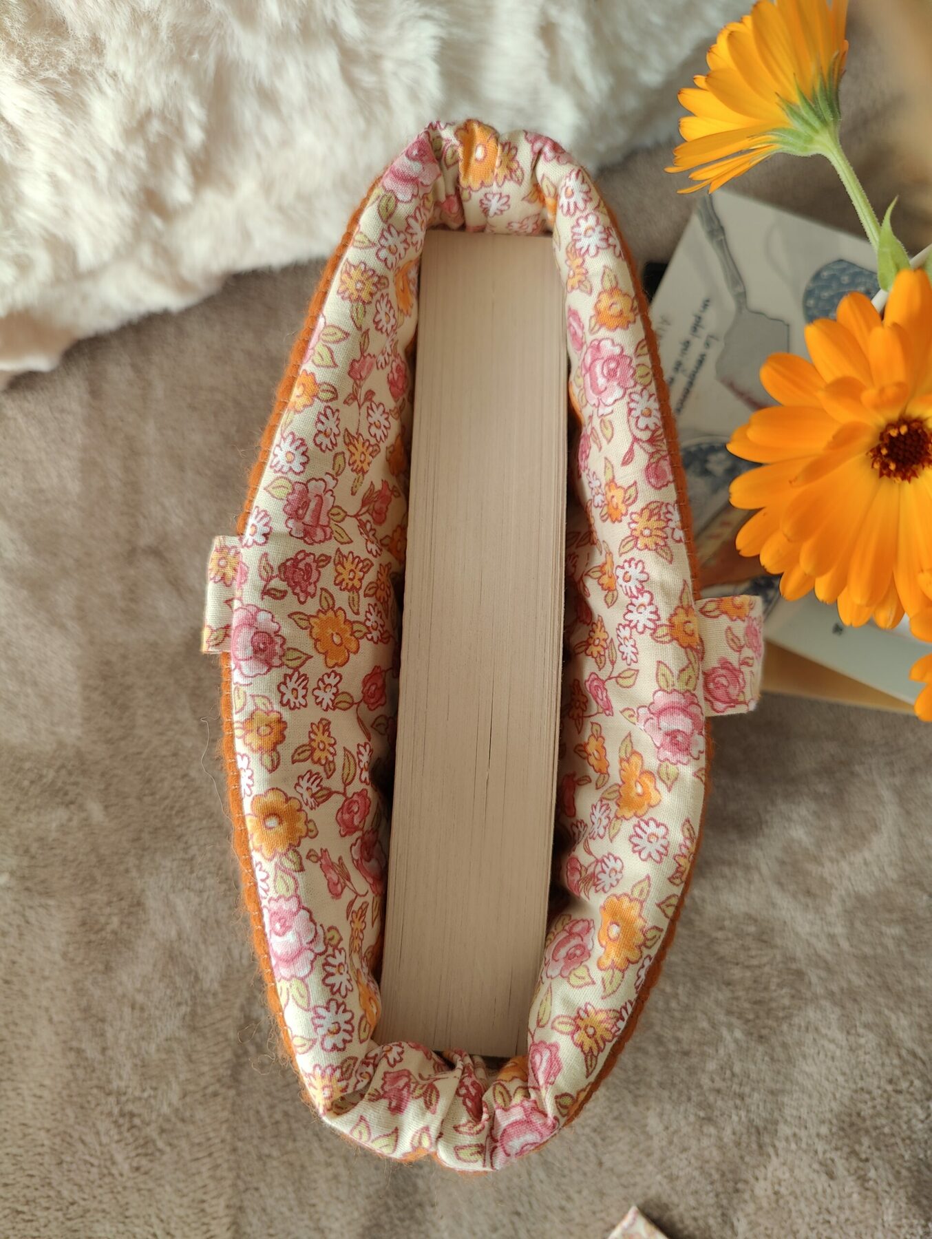 Intérieur fleuri d’une pochette en tissu orangé contenant un livre, sur un lit avec deux oreillers.