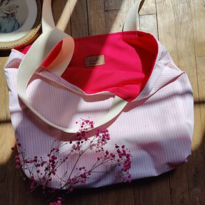 Tote bag réversible upcyclé posé sur un sol en bois, côté rayé blanc et rose visible avec intérieur fuchsia, décoré de gypsophiles roses.